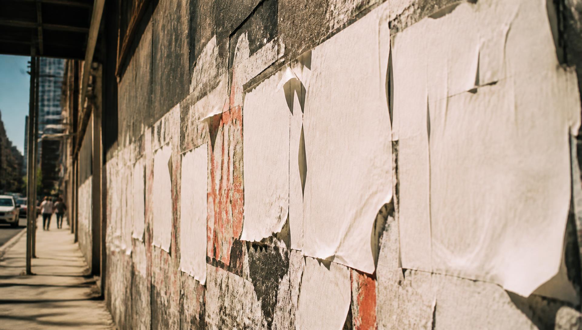 A New York City street wall layered with wheat-pasted posters, torn and weathered in daylight